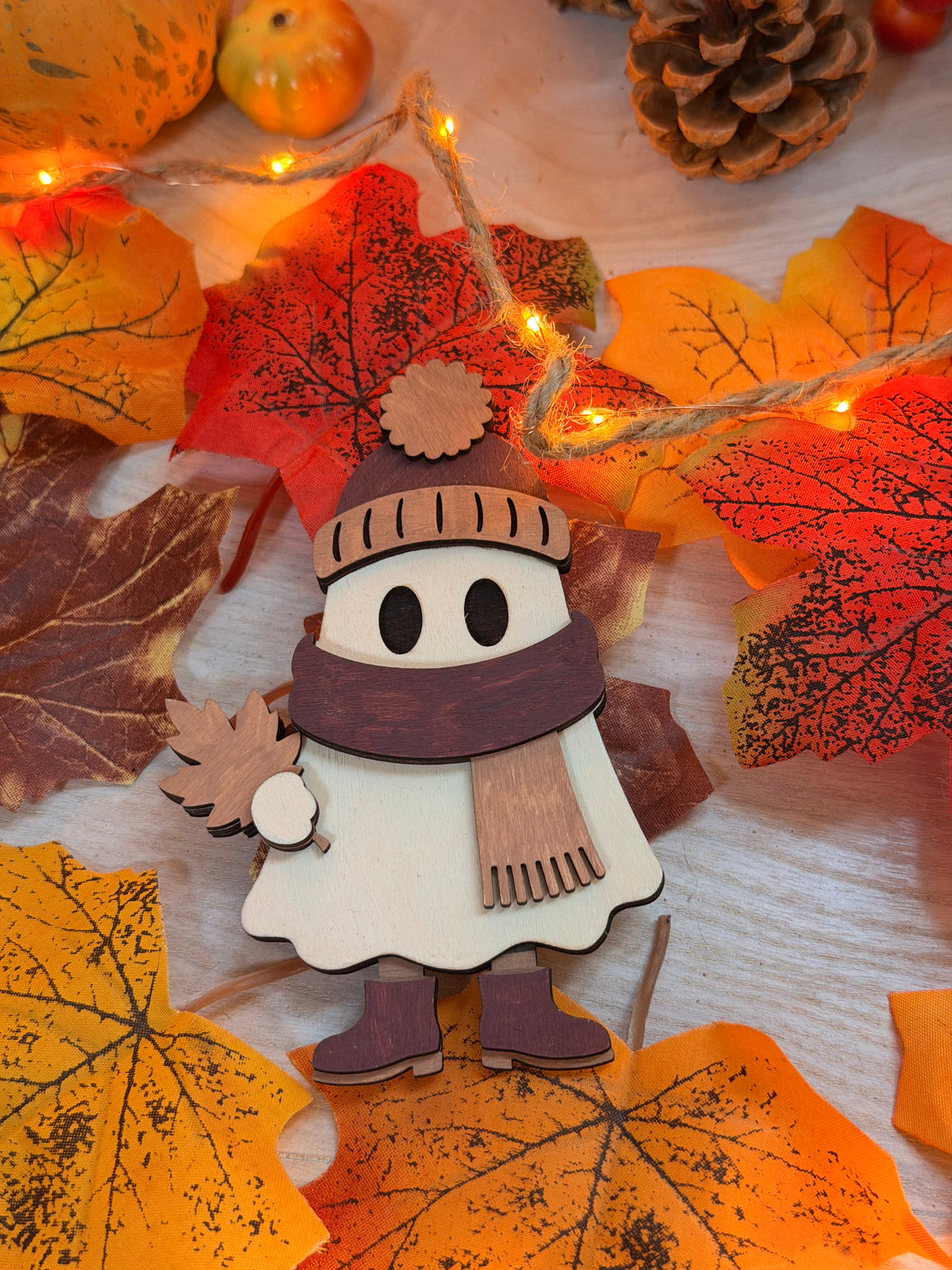 Gaston le Frileux, figurine fantôme en bois découpé au laser et peint à la main avec bonnet, écharpe et bottes, posée sur des feuilles d'automne illuminées - L'Atelier du Moulin