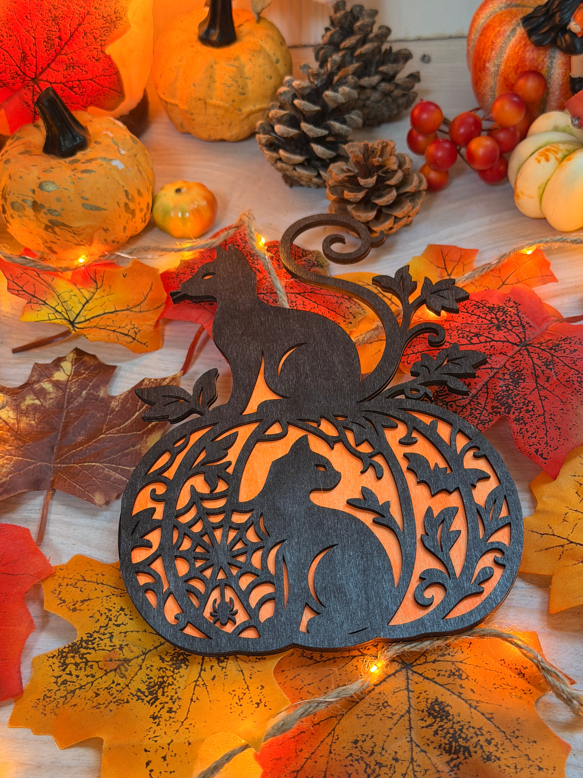 Le Félin d'Automne, décoration en bois noir découpée au laser en forme de citrouille ajourée avec un chat assis au sommet, posée sur des feuilles d'automne illuminées - L'Atelier du Moulin