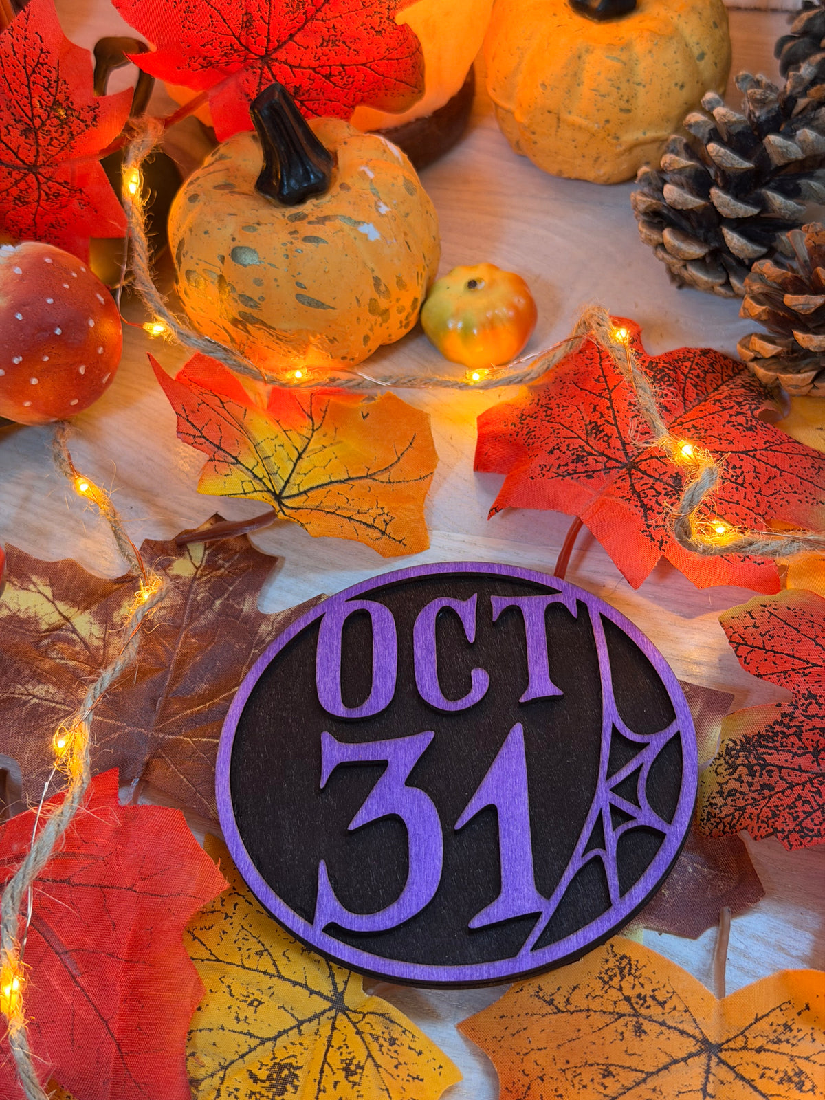 Le Blason d'Halloween, plaque ronde en bois noir et violet gravée au laser "OCT 31" avec toile d'araignée, posée sur des feuilles d'automne illuminées - L'Atelier du Moulin