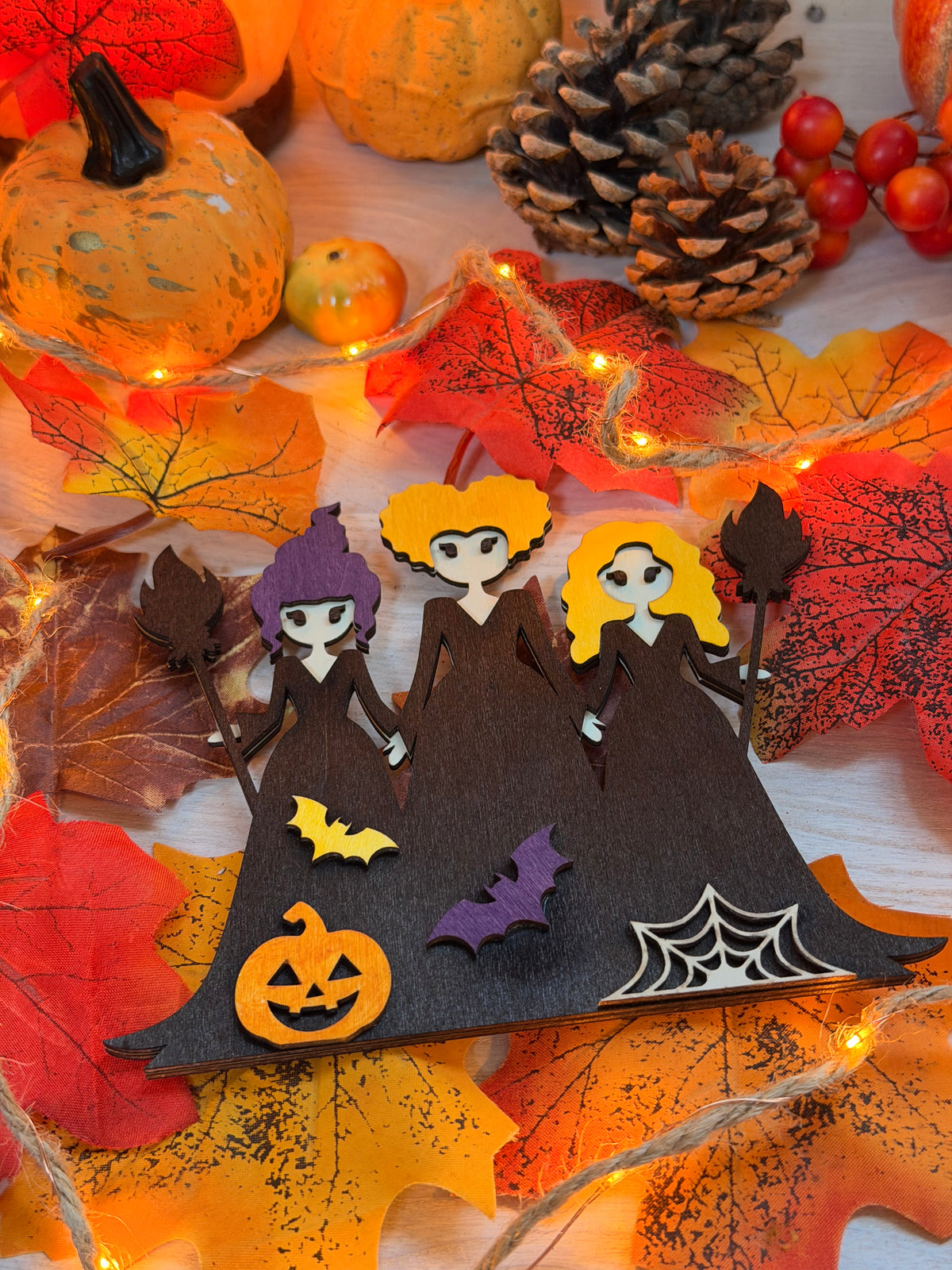 Le Sabbat des Sorcières, silhouette décorative en bois gravé et peint représentant trois sorcières, avec citrouille, chauves-souris et toile d'araignée, posée sur des feuilles d'automne illuminées - L'Atelier du Moulin