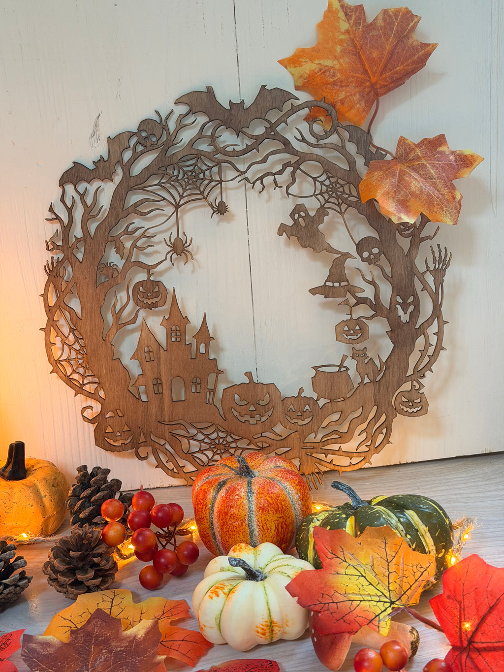 La ronde des Ombres, couronne murale en bois découpée au laser avec scène d'Halloween : maison hantée, chauves-souris, fantômes et citrouilles, posée parmi citrouilles et feuilles d'automne - L'Atelier du Moulin