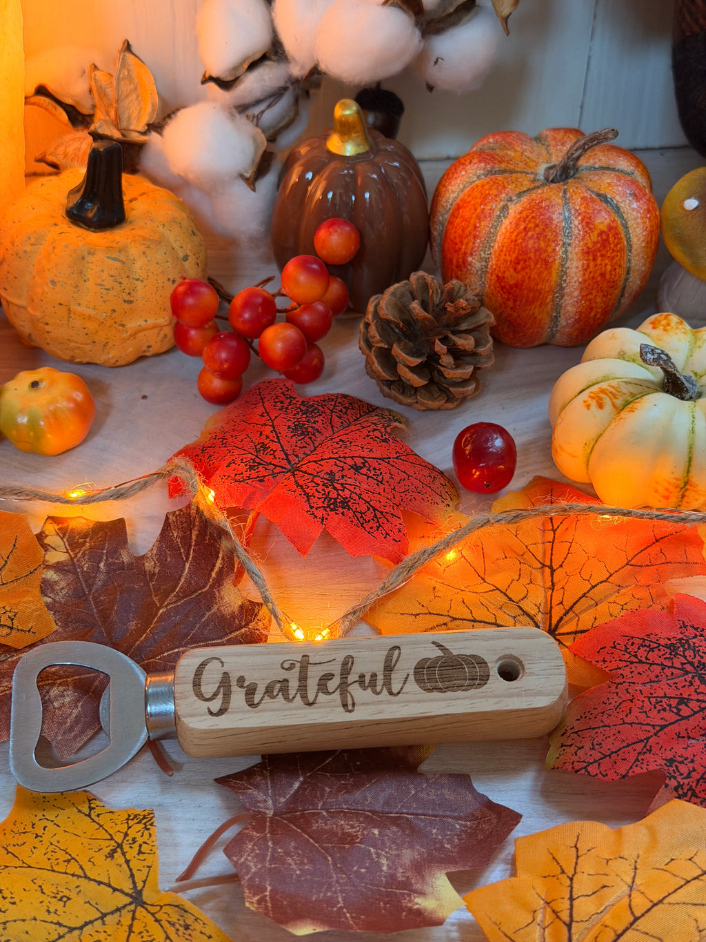 Décapsuleur "Grateful" au manche en bois gravé au laser d'une citrouille, posé sur des feuilles d'automne illuminées parmi citrouilles et pommes de pin - L'Atelier du Moulin