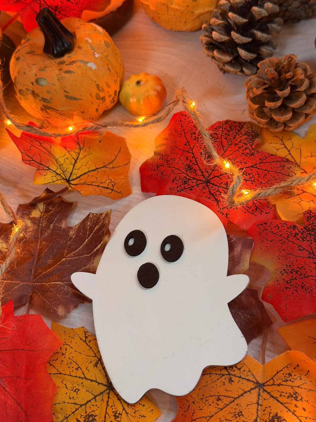Le P'tit Frisson, petit fantôme en bois blanc découpé au laser, posé parmi citrouille, pommes de pin et feuilles d'automne - L'Atelier du Moulin